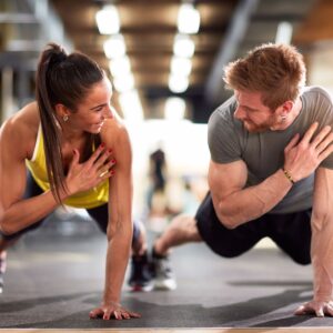 A male and female athlete in a plank position doing shoulder taps while smiling at one another.